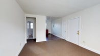 This single-floor space presents a clean, functional layout with a clear separation between living and utility areas. The main living area is finished with wall-to-wall carpet in a neutral brown tone, providing a comfortable underfoot feel, while the adjoining room transitions to dark hardwood flooring, adding contrast and visual interest. Walls throughout are painted in a crisp white, contributing to a bright and open atmosphere despite the modest scale of the space.

A central structural divider partially separates the main living area from the secondary room, which appears to serve as a transitional or dining space. This secondary area features a dark wood cabinet or vanity unit along the wall, alongside what appears to be a radiator, suggesting a traditional heating system is in place. Natural light filters in through windows visible in both the secondary room and the bathroom, keeping the interior from feeling closed off.

A fully enclosed bathroom is accessible directly from the main living area, featuring tile flooring and a window that allows for natural ventilation and light. Two white paneled doors provide entry and interior access, finished with gold-toned hardware. The overall ambiance is modest, clean, and move-in ready.