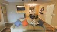 This single-floor residence offers an inviting open-concept design that effortlessly connects the main living areas. The living room serves as the heart of the home, anchored by a plush, light-toned sofa accented with colorful throw pillows in shades of orange, blue, and gray. A ceiling fan overhead adds both function and style to the comfortable space.

The adjoining dining area features a round dark-wood table surrounded by matching chairs, illuminated by a stylish multi-bulb chandelier that adds a warm, intimate ambiance. A large mirror on the dining room wall enhances the sense of depth and reflects light throughout the space.

Flowing naturally from the dining area, the kitchen is defined by striking dark cabinetry that contrasts beautifully against light granite-style countertops. A pass-through counter connects the kitchen to the living and dining spaces, promoting an open and social atmosphere ideal for entertaining.

Warm golden-yellow walls run throughout the shared spaces, creating a cohesive and cozy palette. Decorative floating shelves in the living area provide tasteful display space, while wood-look flooring grounds the entire layout with a clean, modern finish. The overall ambiance is warm, functional, and thoughtfully designed.