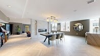 Located at 100 Hale Ave in White Plains, NY, this thoughtfully designed single-floor amenity space offers an open, flowing layout that seamlessly connects a dining area, a fully equipped kitchen, and a vibrant living room. Throughout the space, light-toned herringbone wood flooring provides a cohesive and sophisticated foundation, complemented by clean white ceilings with recessed lighting.

The dining area serves as a central hub, anchored by a bold dark dining table with upholstered gold-accented chairs beneath a striking four-globe pendant light fixture. A floor-to-ceiling concrete accent wall features a linear gas fireplace flanked by decorative stacked log niches, adding warmth and visual drama.

The kitchen is equipped with a substantial stone waterfall island, dark cabinetry, stainless steel appliances, and a textured penny-tile backsplash. Large windows flood this area with abundant natural light and offer views of the surrounding White Plains skyline.

The living room is designed for entertainment, featuring a pool table beneath a Tiffany-style pendant light, a tufted leather sofa, velvet accent chairs, built-in shelving, a wall-mounted television, and classic arcade machines. The earthy olive-toned accent walls and layered area rugs create an inviting, lounge-like atmosphere throughout this dynamic and well-appointed space.
