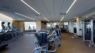 This fitness center offers a well-appointed workout environment designed to accommodate a wide range of exercise needs. The space is laid out on a single floor, with a thoughtful arrangement that allows for easy navigation between equipment zones. Overhead, a suspended ceiling features striking linear LED lighting strips that run the length of the room, casting bright, even illumination throughout the facility.

The strength training area is prominently featured, housing a robust selection of cable machines, resistance equipment, and weight stations. A dedicated free weights section lines one wall, with a neatly organized dumbbell rack visible in the background. The cardio zone is positioned toward the perimeter of the space, with treadmills and additional cardio machines placed near large windows that allow natural light to filter in and provide exterior views.

Multiple flat-screen televisions are mounted throughout the facility, offering entertainment during workouts. The flooring transitions between rubber and hard surfaces, delineating functional zones. The overall ambiance is clean, modern, and professional, reflecting a well-maintained fitness facility suited for a variety of training routines.