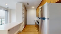 Located at 1028 Stitch Bend Way in Charlotte, NC, this efficiently designed single-floor unit makes smart use of its intimate footprint. Light hardwood flooring runs continuously throughout the space, creating a cohesive and warm aesthetic.

The galley-style kitchen is well-appointed with warm wood upper cabinetry, stainless steel appliances including a full-size refrigerator and range, and a clean white countertop. A tall built-in shelving unit along one wall provides generous open storage and organizational versatility.

The living area is bright and airy, distinguished by a full-height sliding glass door that opens to a private balcony, offering views of the surrounding Charlotte neighborhood. A sidelight window further enhances natural light in this open space, contributing to an inviting atmosphere.

The bathroom is finished in a clean, contemporary style with light wood-look flooring, a white vanity with undermount sink, a dark-framed mirror, and matte black hardware and fixtures. White wall tiles in the tub surround area reinforce the crisp, modern aesthetic.

Overall, the unit presents a practical yet polished design, well-suited for those seeking a low-maintenance, modern living space in Charlotte's developing urban landscape.