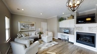 This single-story space at Maple Brook offers a well-designed open layout that seamlessly blends comfort and functionality. The main living area features warm hardwood-style flooring, neutral gray-toned walls, and crown molding that adds a refined architectural detail. Recessed lighting throughout the ceiling ensures consistent illumination, complemented by a decorative orb-style chandelier that serves as a subtle focal point.

The living area is generously sized, accommodating a full seating arrangement with plush sofas and accent chairs arranged around a central area rug with a geometric pattern. A large framed landscape artwork anchors one wall, contributing to the calm, cohesive aesthetic of the space.

A standout feature is the built-in workstation along one wall, complete with white cabinetry above and below the desk surface, providing ample storage. A textured stone tile backsplash behind the workspace adds visual interest and contrast against the white cabinetry. Decorative greenery atop the upper cabinets introduces a natural element. The overall ambiance is bright, clean, and practical, making the space suitable for both relaxing and working.