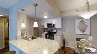 This single-floor space offers a thoughtfully designed layout centered around a stylish, open-concept kitchen and dining area. The kitchen is a functional and visually appealing space, featuring stainless steel appliances including a refrigerator, electric range, and built-in microwave. White cabinetry provides ample storage, while the light granite countertops add a touch of elegance and contrast beautifully against the clean, neutral color palette throughout. A curved peninsula with an undermount sink extends the prep and seating area, enhanced by two pendant lights with classic bell-shaped shades that cast warm, focused illumination over the workspace. The textured ceiling adds subtle architectural character to the kitchen area. Adjacent to the kitchen, a compact dining space accommodates a glass-top table and chairs, creating a convenient area for meals. The warm-toned flooring runs throughout, contributing to a cohesive and inviting ambiance. Overall, the space balances practicality with modern design, offering a comfortable and well-equipped environment suited for everyday living.
