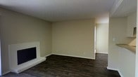 This single-story space presents a straightforward and functional layout centered around a living area that serves as the heart of the interior. The room is finished with dark hardwood-style plank flooring, which adds a sense of warmth and contrast against the neutral beige-toned walls. A textured popcorn ceiling runs overhead, a characteristic detail of the space's construction style.

A notable feature of the living area is the fireplace positioned along one wall, framed by a clean, light-colored surround and raised hearth that provides a simple focal point. The walls are left largely open, offering flexibility for furniture arrangement and personalization.

An adjacent area is visible through an open passageway, suggesting a connected layout that allows for natural flow between spaces. Standard electrical outlets are distributed along the walls, and white baseboards provide a clean transition between the flooring and wall surfaces. The overall ambiance is understated and neutral, presenting a blank canvas that is well-suited for a variety of interior design approaches.
