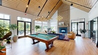 Located on Buckingham Street in Matawan, NJ, this thoughtfully designed single-floor space offers a generous and versatile layout across its expansive footprint. The centerpiece is a dramatic great room boasting soaring vaulted ceilings clad in warm wood paneling, illuminated by sculptural gold chandelier fixtures that cast an inviting glow over light oak hardwood floors. A full-height brick fireplace anchors the living area, while large black-framed windows and glass double doors flood the space with natural light and offer views of the outdoor patio and surrounding greenery. A regulation pool table adds a recreational focal point to the open living area. The kitchen features sleek slate-blue cabinetry, white quartz countertops, a deep undermount sink, and a striking geometric metallic tile backsplash, complemented by a sliding glass door leading to the exterior. The dining room presents an impressive marble-top table beneath warm pendant lighting, set against sage-green walls with glass-paneled sliding doors. A separate private dining/meeting room provides an intimate setting with a dark wood table, leather chairs, and built-in cabinetry with a wall-mounted display. The welcoming entry foyer transitions seamlessly between areas, featuring tile flooring and built-in shelving, completing this well-appointed and communal space.