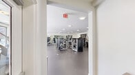 Located at 527 Old Bridge Tpke in East Brunswick, NJ, this two-floor fitness facility offers a comprehensive and well-appointed space designed to support a wide range of exercise activities. The layout spans two generously sized floors connected by staircases, providing ample room for both strength training and group fitness pursuits.

The main exercise floor is outfitted with an extensive array of Life Fitness strength machines and cable systems, arranged across durable rubberized flooring that defines the space's functional aesthetic. Large floor-to-ceiling windows along one wall flood the cardio zone with natural light while offering pleasant outdoor views, creating an energizing environment for treadmill and elliptical workouts. Wall-mounted mirrors throughout the strength area enhance the sense of openness and allow for proper form monitoring.

The second exercise area serves as a dedicated multi-purpose studio, featuring rich hardwood flooring, a wall-mounted ballet barre, and mirrored walls. This versatile space accommodates yoga, stretching, cycling, and group classes, with equipment including spin bikes and medicine ball racks neatly organized along the perimeter. Interior windows connect this studio to adjacent spaces, contributing to an open, integrated feel.

Throughout both floors, recessed and flush-mount ceiling lighting ensures consistent, bright illumination. The overall design is clean, modern, and functional, providing a professional-grade fitness environment.