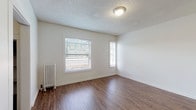 Located on SW Montgomery Street in Portland, Oregon, this single-floor unit offers a clean, functional layout with consistent finishes throughout. Rich wood-style plank flooring runs continuously across all rooms, providing a warm, cohesive aesthetic that contrasts cleanly with the bright white walls found throughout the space.

The living areas are well-lit, benefiting from multiple windows fitted with horizontal blinds that allow for adjustable natural light. Textured ceilings and flush-mount light fixtures are present in several rooms, contributing to a classic residential character. Cast-iron radiators positioned along the walls serve as both a functional heating source and a period-appropriate architectural detail.

The kitchen is equipped with white shaker-style cabinetry, butcher block countertops, an apron-front stainless steel sink, open wall shelving, and a subway tile backsplash — offering a practical yet stylish workspace. The bathroom features a tiled tub-shower surround, a white vanity with drawers, and matching wood-style flooring.

Additional rooms provide versatile living or sleeping spaces, each featuring natural light from surrounding windows, including steel-framed windows with views of neighboring brick architecture. A hallway entry with a bold navy blue door provides a distinct entryway into the unit.