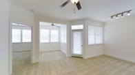 This single-floor space offers a clean, light-filled interior defined by a neutral color palette and practical design choices throughout. The living area is finished with plush wall-to-wall carpeting in a warm beige tone, contributing to a comfortable and welcoming atmosphere. Crisp white walls and white trim create a bright, airy feel that is further enhanced by multiple windows dressed with horizontal blinds, allowing for flexible light control while drawing in generous amounts of natural light.

Ceiling fans are present in the main living areas, providing both functional air circulation and subtle visual interest overhead. Track lighting is also installed along the ceiling, offering directed illumination to complement the natural light. A paneled interior door frames the entry into the primary living space, adding a classic architectural touch.

A secondary area — visible through an open archway — mirrors the main room's finishes, featuring its own ceiling fan and additional windows that wrap the space in light. This open-concept layout connects the areas fluidly, making the overall space feel expansive and versatile. An exterior door with a transom window above provides direct access and contributes to the room's brightness. The overall ambiance is clean, move-in ready, and well-suited to a variety of furnishing styles.