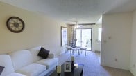 This single-floor residence offers a welcoming and functional layout designed for comfortable everyday living. The open-plan living and dining area serves as the heart of the space, where soft wall-to-wall carpeting adds warmth underfoot and a textured ceiling lends a classic finish overhead. A ceiling fan with an integrated light fixture provides both air circulation and ambient lighting to the combined area.

The living area is generously proportioned, offering ample room for seating arrangements centered around a dark coffee table, creating an inviting and relaxed atmosphere. Adjacent to the living space, the dining area accommodates a glass-top table with wrought iron chairs, positioned to take full advantage of natural light streaming in through a large sliding glass door. This door opens directly onto a balcony, providing outdoor views and enhancing the sense of openness throughout the interior.

A framed wall piece adds a decorative focal point between the living and dining zones, contributing to the overall cohesive aesthetic. Natural light flows generously through the sliding door and an additional window nearby, keeping the interior bright and airy. A glimpse of an adjoining room suggests further functional spaces within the layout, rounding out this well-appointed single-level home.