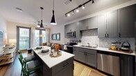 This single-floor residence showcases a thoughtfully designed open-concept layout that seamlessly connects the kitchen, dining, and living areas. The kitchen is a standout feature, equipped with grey shaker-style cabinetry extending from floor to ceiling, a herringbone-patterned marble tile backsplash, and a full suite of stainless steel appliances including an electric range, over-the-range microwave, dishwasher, and French door refrigerator. White quartz countertops extend across the perimeter and onto a central island, which provides additional seating and prep space beneath striking black cone-shaped pendant lights. Track lighting overhead ensures the kitchen remains well-lit throughout. Hardwood-style flooring runs throughout the main living areas, adding warmth and continuity. The living area benefits from natural light streaming through large windows dressed with sheer curtains, and a ceiling fan provides additional comfort. The bedroom is a calm, private retreat featuring hardwood-style flooring, a window with exterior views, and ample space for furnishings. The bathroom includes a full soaking tub and shower combination with a curved curtain rod, grey vanity cabinetry, and quartz countertops, maintaining the cohesive modern aesthetic found throughout the residence.