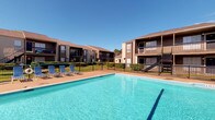 This outdoor pool area serves as a central amenity within a well-established residential community, offering a refreshing retreat under open skies. The generously sized swimming pool features clear, blue water and is framed by a broad pool deck finished in a warm-toned concrete surface, providing ample space for sunbathing and relaxation. Several lounge chairs are positioned poolside, offering comfortable spots to unwind in the sun. The pool is enclosed by a sleek black metal safety fence, ensuring both security and an unobstructed visual connection to the surrounding grounds. Neatly trimmed hedges and mature landscaping line the perimeter, adding a layer of greenery that enhances the overall atmosphere. The backdrop consists of two-story residential buildings with brown and tan exteriors, wood-accented balconies, and exterior staircases that contribute to the community's cohesive architectural character. The open layout of the space allows for abundant natural light throughout the day, and the clear blue sky overhead reinforces the bright, welcoming ambiance. This pool area is well-suited for both active recreation and leisurely outdoor enjoyment, making it a standout feature of the community.