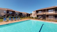 This outdoor pool area serves as a central amenity within a well-established residential community, offering a refreshing retreat under open skies. The generously sized swimming pool features clear, blue water and is framed by a broad pool deck finished in a warm-toned concrete surface, providing ample space for sunbathing and relaxation. Several lounge chairs are positioned poolside, offering comfortable spots to unwind in the sun. The pool is enclosed by a sleek black metal safety fence, ensuring both security and an unobstructed visual connection to the surrounding grounds. Neatly trimmed hedges and mature landscaping line the perimeter, adding a layer of greenery that enhances the overall atmosphere. The backdrop consists of two-story residential buildings with brown and tan exteriors, wood-accented balconies, and exterior staircases that contribute to the community's cohesive architectural character. The open layout of the space allows for abundant natural light throughout the day, and the clear blue sky overhead reinforces the bright, welcoming ambiance. This pool area is well-suited for both active recreation and leisurely outdoor enjoyment, making it a standout feature of the community.