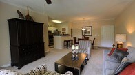 This single-floor residence showcases an open-concept layout that connects the living room, dining area, and kitchen into one cohesive, flowing space. The living room features plush carpeting, crown molding, and a ceiling fan, and is furnished with patterned upholstered seating arranged around a dark wood coffee table. A large dark wood entertainment armoire anchors one wall, adding a bold contrast to the neutral, warm-toned walls.

The dining area sits adjacent to the living room and is defined by an elegant chandelier hanging above a dark wood dining table with upholstered chairs. A framed piece of wall art complements the space, adding a decorative touch.

The kitchen is partially open to the main living area via a peninsula breakfast bar with bar stool seating. It features stainless steel appliances, including a refrigerator and range, set against light cabinetry. Tile flooring in the kitchen contrasts with the carpeted living and dining areas. The overall ambiance of the space is warm and inviting, with consistent crown molding throughout reinforcing a polished, cohesive aesthetic.