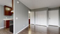This single-floor unit offers a well-organized layout designed for practical, comfortable living. Throughout the space, durable hardwood-style flooring in warm tones provides a cohesive foundation, complemented by cool gray walls and crisp white trim and baseboards that give the interior a clean, modern feel.

The open living area is bright and inviting, featuring a large window dressed with vertical blinds that allows natural light to fill the room while offering views of the surrounding greenery. A baseboard heating unit and wall-mounted air conditioner ensure year-round climate comfort.

The kitchen is efficiently arranged in a galley-style configuration, featuring rich dark wood cabinetry both above and below the counter, light-toned countertops with an integrated sink, and black appliances including an electric range with overhead ventilation hood and a stainless steel refrigerator.

The bedroom is generously sized and includes expansive double bi-fold closets offering substantial storage. A doorway connects directly to the bathroom, which is fitted with a full-size soaking tub and shower combination, a wood-toned vanity, and hardwood-style flooring that ties back to the rest of the unit.

The space also features a textured ceiling treatment and a front entry door opening directly into the main living area, completing this functional and neatly finished single-floor home.
