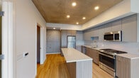 Located in the heart of Portland's vibrant West End neighborhood, this single-floor residence at 1510 SW Alder St offers a sleek, modern aesthetic with high-end finishes throughout. The open-concept kitchen serves as the centerpiece of the space, showcasing a large central island with a crisp white quartz countertop, ideal for dining or meal preparation. The kitchen is outfitted with a full suite of stainless steel appliances, including a gas range, built-in microwave, dishwasher, and refrigerator. Two-tone cabinetry — warm wood lower cabinets paired with matte gray upper cabinets — adds visual depth, while a delicate subway tile backsplash provides a refined finishing touch. Striking exposed concrete ceilings lend an industrial character to the space, contrasting beautifully with the warm-toned hardwood flooring that flows throughout the main living areas. The bathroom features floor-to-ceiling marble tile surrounding the soaking tub and shower, paired with a wood-grain vanity and clean white countertop for a spa-like feel. A generously sized walk-in closet with built-in wire shelving provides ample storage. Recessed lighting throughout ensures the space is well-lit, creating a bright and welcoming ambiance.