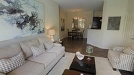 This single-floor residence offers a thoughtfully designed open-concept layout that connects the living, dining, and kitchen areas in a cohesive and functional flow. The living area is anchored by rich, dark hardwood flooring and neutral-toned walls that create a warm, inviting ambiance. A plush sofa with patterned accent pillows faces a sleek dark coffee table, complemented by a large abstract canvas artwork that adds a contemporary artistic touch to the space. A soft area rug defines the seating zone, adding texture and comfort underfoot. The space flows naturally into the dining area, where a dark wood dining table and matching chairs are centered beneath an elegant chandelier, lending a refined character to everyday gatherings. A decorative mirror on the dining room wall enhances the sense of depth and light in the space. Just beyond, the kitchen is accessible via a pass-through counter, offering a practical and visually open connection to the main living areas. Dark cabinetry in the kitchen provides a bold contrast to the lighter tones throughout the rest of the home. Overall, the space strikes a balance between modern sophistication and comfortable, livable design.
