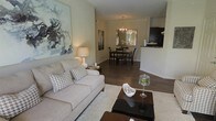 This single-floor residence offers a thoughtfully designed open-concept layout that connects the living, dining, and kitchen areas in a cohesive and functional flow. The living area is anchored by rich, dark hardwood flooring and neutral-toned walls that create a warm, inviting ambiance. A plush sofa with patterned accent pillows faces a sleek dark coffee table, complemented by a large abstract canvas artwork that adds a contemporary artistic touch to the space. A soft area rug defines the seating zone, adding texture and comfort underfoot. The space flows naturally into the dining area, where a dark wood dining table and matching chairs are centered beneath an elegant chandelier, lending a refined character to everyday gatherings. A decorative mirror on the dining room wall enhances the sense of depth and light in the space. Just beyond, the kitchen is accessible via a pass-through counter, offering a practical and visually open connection to the main living areas. Dark cabinetry in the kitchen provides a bold contrast to the lighter tones throughout the rest of the home. Overall, the space strikes a balance between modern sophistication and comfortable, livable design.