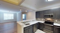 This single-floor residence showcases a thoughtfully designed open-concept layout that connects the kitchen and living areas in a seamless, functional flow. The kitchen is a standout feature, equipped with dark espresso-toned cabinetry that provides generous storage, complemented by crisp white countertops that offer a striking contrast. A peninsula with an undermount sink adds both workspace and a natural dividing point between the kitchen and the adjoining living space. Stainless steel appliances, including a range, microwave, and dishwasher, lend a modern, polished look throughout. A decorative mosaic tile backsplash in neutral tones adds texture and visual interest to the cooking area. Track lighting illuminates the kitchen, while recessed tray ceilings with crown molding detailing elevate the overall aesthetic. The transition into the living area reveals warm hardwood-style flooring and an arched entryway that adds architectural character. The living space is bathed in natural light from a large window, creating a bright and welcoming ambiance. Light tile flooring in the kitchen transitions gracefully to the warmer tones of the adjoining room, tying the design together cohesively throughout the home.