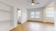 This efficiently designed one-bedroom, one-bathroom unit makes thoughtful use of its compact footprint across a single floor. Light wood flooring runs throughout, creating a cohesive and clean aesthetic that enhances the sense of openness in every room.

The combined kitchen and living area offers a functional layout. The kitchen features gray cabinetry, white subway tile backsplash, light countertops, and a full suite of stainless steel appliances including a gas range, microwave, and refrigerator. Baseboard heating is present throughout the unit, and multiple windows allow natural light to brighten the living spaces.

The bedroom includes built-in open shelving along one wall, providing convenient storage and display space, as well as access to a dedicated closet with a hanging rod. A ceiling fan serves the main living area for added comfort.

The bathroom is finished in white tile and features a pedestal sink, a mirror, and a full soaking tub with a shower. A window provides natural ventilation. The overall ambiance of the unit is clean, bright, and straightforward, with a neutral white palette and warm wood floors throughout.