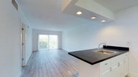 This single-floor space showcases a clean, modern design with an open-concept layout that seamlessly connects the kitchen and living areas. Light wood-toned flooring runs throughout, lending warmth and continuity to the space, while crisp white walls and recessed lighting create a bright, airy atmosphere.

The kitchen is a standout feature, anchored by a peninsula with a dark countertop that provides both prep space and a casual seating area. White shaker-style cabinetry offers ample storage, and a dark tile backsplash adds a contrasting design detail. Stainless steel appliances — including a refrigerator, electric range, and range hood — complete the well-equipped cooking area.

The living area flows naturally from the kitchen, benefiting from an abundance of natural light streaming in through large sliding glass doors fitted with vertical blinds. These doors open to an outdoor space and frame views of lush greenery, bringing the outside in and enhancing the overall sense of openness.

The layout also includes a closet with louvered doors and a separate entry door, offering practical storage solutions. A stepped ceiling detail above the kitchen zone adds architectural interest. The overall ambiance is light, clean, and contemporary — a well-designed space that balances functionality with modern aesthetics.