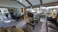This single-floor space showcases a thoughtfully designed open-concept layout that seamlessly integrates the living, dining, and kitchen areas into one cohesive environment. The vaulted ceiling with exposed dark wooden beams adds striking architectural character, while dual ceiling fans ensure comfortable airflow throughout. Large screened windows span the perimeter of the living area, allowing abundant natural light to pour in and offering views of the surrounding greenery outdoors.

The living area is anchored by a rich, dark wood coffee table set atop a plush area rug, complemented by patterned accent chairs and a leather sofa — creating an inviting and well-appointed seating arrangement. Adjacent to the living space, the dining area features a substantial dark wood table with matching chairs, suitable for hosting gatherings. A glimpse into the kitchen reveals white cabinetry and pendant lighting, hinting at a functional and well-lit workspace.

Terracotta-toned tile flooring runs throughout, adding warmth and cohesion to the space. A wooden storage cabinet serves as a decorative focal point near the screened wall, enhancing the room's earthy, relaxed aesthetic. Overall, this space combines casual comfort with architectural detail to create an open, airy, and welcoming atmosphere.