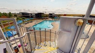 Located at 399 S Trussville St in Aurora, Colorado, Splash Park is a comprehensive outdoor aquatic facility designed to offer recreational water experiences for all ages. The park's centerpiece is a large, open lap pool with a freeform leisure section, surrounded by a generously sized concrete deck lined with lounge chairs and colorful umbrellas in vivid blues and oranges. A prominent multi-level water slide tower anchors one corner of the park, featuring a winding staircase and multiple slide options, including a covered blue water slide with sweeping views of the surrounding Colorado plains. Adjacent to the slide tower is a dedicated splash pad area equipped with interactive water jets and spray features scaled for younger visitors. A circular hot tub with blue tile accents is nestled within a landscaped greenspace, offering a relaxing counterpoint to the more active water attractions. The park's support building features a modern design with wood-panel cladding and stone accents, housing restrooms and additional amenities. A covered outdoor lounge area with seating provides a comfortable gathering space, while mature trees and manicured landscaping soften the perimeter. The facility enjoys expansive open-sky views characteristic of the Colorado Front Range.