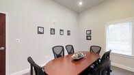 This single-floor conference space offers a clean, professional environment well-suited for focused meetings and collaborative discussions. The room centers around a warm cherry-toned oval conference table surrounded by black mesh ergonomic chairs, providing comfortable seating for multiple attendees. The walls are finished in a neutral off-white palette that creates a bright, calm atmosphere, complemented by recessed ceiling lighting that ensures even, consistent illumination throughout the space. A notable design element is the gallery wall arrangement of black-and-white framed photographs, lending the room a distinguished and personalized character that sets it apart from a standard meeting room. The decor is kept minimal and purposeful — a decorative bowl centerpiece on the table adds a subtle touch of warmth without distracting from the room's professional function. The space benefits from clean architectural lines, white baseboards, and a well-proportioned layout that feels both intimate and functional. Overall, the room strikes a balance between a polished corporate aesthetic and an approachable, welcoming ambiance, making it an ideal setting for small group meetings, presentations, or private discussions.