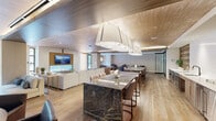 Located on Atlas Street in Baltimore's 21230 zip code, this single-floor amenity space combines refined design with functional versatility. The interior is unified by warm wood-plank flooring and striking wood-paneled ceilings that lend a cohesive, contemporary aesthetic throughout. A central gathering area anchors the space with two substantial dark marble-topped islands supported by brushed gold bases, surrounded by cushioned dining chairs—ideal for communal dining or social events. Overhead, a dramatic sculptural pendant light fixture commands attention while providing generous illumination across the space.

The kitchen area is well-appointed with stainless steel appliances, a dishwasher, oven, and refrigerators, alongside clean white countertops and light wood cabinetry. Distinctive decorative wall panels featuring circular cutout patterns flank the kitchen, adding an artistic architectural element. A generous lounge area offers a large sectional sofa oriented toward a wall-mounted flat-screen television set within a warm wood surround, creating an inviting media and relaxation zone. A separate seating lounge near glass entry doors allows natural light to fill the space. Additional areas visible through glass partitions include a fitness room. The overall ambiance is polished, warm, and thoughtfully designed for community use.