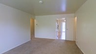 This single-floor space presents a clean, straightforward layout designed for functional everyday living. The main living area is finished with wall-to-wall beige carpeting, offering a comfortable underfoot feel, and is framed by smooth white walls that create a bright, neutral backdrop. Standard-height ceilings keep the space feeling tidy and manageable, while a smoke detector is visibly installed overhead.

An open doorway connects the carpeted living area to an adjacent room featuring hard flooring—likely a kitchen or dining area—which benefits from notable natural light streaming in through what appears to be a sliding glass door or large exterior door. This transition between flooring materials helps visually define the two functional zones.

A hallway is accessible from the main living space, suggesting additional rooms beyond the primary area. A closet door is also visible along one wall, providing convenient storage. The overall ambiance is clean and move-in ready, with a simple, unfurnished interior that allows for flexible arrangement. The abundance of natural light visible in the adjoining area contributes to an open, airy feel throughout the space.