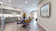 This single-floor residence features a thoughtfully designed open-concept layout that seamlessly connects the kitchen, dining area, and living room. Light wood-style flooring flows throughout the main living spaces, complemented by neutral-toned walls and recessed lighting that create a bright, welcoming atmosphere.

The kitchen is outfitted with gray shaker-style cabinetry extending to the upper walls, light granite countertops, a white subway tile backsplash, and stainless steel appliances including a range, microwave, and dishwasher. The adjacent dining area accommodates a round table with stylish upholstered seating, while a decorative wall-mounted mirror adds visual depth near the entry.

The living room benefits from a full-length glass door that invites natural light, with ample space for comfortable seating arrangements. The bedroom features plush carpeting, a ceiling fixture, and a window for natural light. A well-proportioned walk-in closet with built-in shelving and hanging rods provides generous storage capacity.

The bathroom is finished with wood-look tile flooring, a walk-in shower with white subway tile surround, a toilet, and a gray vanity with a granite countertop, maintaining the cohesive, contemporary aesthetic found throughout the residence.