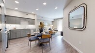 This single-floor residence features a thoughtfully designed open-concept layout that seamlessly connects the kitchen, dining area, and living room. Light wood-style flooring flows throughout the main living spaces, complemented by neutral-toned walls and recessed lighting that create a bright, welcoming atmosphere.

The kitchen is outfitted with gray shaker-style cabinetry extending to the upper walls, light granite countertops, a white subway tile backsplash, and stainless steel appliances including a range, microwave, and dishwasher. The adjacent dining area accommodates a round table with stylish upholstered seating, while a decorative wall-mounted mirror adds visual depth near the entry.

The living room benefits from a full-length glass door that invites natural light, with ample space for comfortable seating arrangements. The bedroom features plush carpeting, a ceiling fixture, and a window for natural light. A well-proportioned walk-in closet with built-in shelving and hanging rods provides generous storage capacity.

The bathroom is finished with wood-look tile flooring, a walk-in shower with white subway tile surround, a toilet, and a gray vanity with a granite countertop, maintaining the cohesive, contemporary aesthetic found throughout the residence.