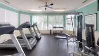 Located at 8905 Poppy Hills Ln in Charlotte, NC, this well-appointed fitness center offers a comprehensive workout environment designed for both cardio and strength training. The single-floor space is finished with soft mint-green walls accented by classic white wainscoting and crown molding, creating a clean and welcoming atmosphere. Durable rubber flooring covers the entire area, providing a safe and functional surface throughout. Large windows fitted with white horizontal blinds line the walls, allowing generous natural light to fill the room while offering views of the surrounding greenery. Ceiling fans ensure comfortable air circulation during workouts. The cardio section features multiple treadmills positioned to take advantage of the natural light, while the strength training area is equipped with a multi-station cable machine, a Magnum weight system, a free-weight bench, and a rack of dumbbells in varying sizes. Full-length mirrors are mounted along one wall, expanding the visual space and allowing users to monitor their form. A wall-mounted television provides entertainment, and framed exercise reference charts are displayed for guidance. The space is well-lit with recessed and flush-mount lighting fixtures, and direct exterior access is provided through glass-panel doors, offering easy entry and exit.