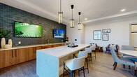 Located at 4805 Creek Cir in Cumming, GA, this thoughtfully designed community lounge offers a refined and welcoming atmosphere ideal for gathering and relaxing. The open single-floor layout is anchored by a striking central island topped with a light-veined quartz surface and flanked by upholstered bar seating, with a trio of geometric pendant lights suspended above. Along one wall, warm wood cabinetry with dark hardware supports a quartz countertop with an integrated sink, all set against a bold dark-green glazed brick tile backsplash that extends to the ceiling. Dual flat-screen TVs are mounted on this feature wall, enhancing the lounge's entertainment appeal. Wood-look tile flooring flows throughout, complementing both the natural warmth of the cabinetry and the cooler tones of the furnishings. Seating areas are thoughtfully arranged with upholstered accent chairs gathered around round side tables, backed by a warm slatted wood panel accent wall that adds texture and depth. Floor-to-ceiling windows and a glass door flood the space with natural light while framing views of the landscaped courtyard and surrounding community. The overall design blends contemporary style with functional comfort, creating an inviting communal environment.