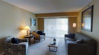 This single-floor residential space offers a well-appointed living area designed for both comfort and functionality. The living room serves as the heart of the space, featuring warm, neutral-toned walls that create an inviting atmosphere. A standout element is the expansive floor-to-ceiling sliding glass door dressed with vertical blinds, which floods the room with abundant natural light and provides direct access to an outdoor balcony with views of the surrounding greenery.

The living area is generously sized, accommodating a comfortable seating arrangement that includes a sofa, an accent armchair, and a sleek coffee table — all centered around a layout ideal for relaxing or entertaining. Soft carpeted flooring adds warmth underfoot and contributes to the cozy ambiance of the room. Tasteful wall art and coordinated furnishings enhance the overall aesthetic.

Ambient lighting from table lamps and floor lamps complements the natural light, ensuring the space remains bright and welcoming at any time of day. The clean lines and practical layout of this single-level residence make it an efficient and comfortable living environment, with easy flow and thoughtful use of available space throughout.