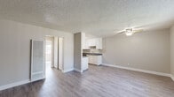 This single-story unit in Suisun City, California (94585) features a neutral, modern aesthetic carried consistently throughout its rooms. Light gray wood-style flooring runs throughout the space, complementing the soft beige and gray-toned walls found in every room.

The open living area is anchored by a textured popcorn ceiling and benefits from natural light streaming through large windows fitted with vertical blinds. A wall-mounted air conditioning unit provides climate control, and a ceiling fan adds additional airflow to the space. The layout flows naturally toward the kitchen, which is partially separated by a half-wall partition. The kitchen features white cabinetry, dark speckled countertops, a stainless steel refrigerator, and a dishwasher, offering practical functionality.

The bedroom area connects to a bathroom that includes a vanity with white cabinetry, a mirror medicine cabinet with overhead lighting, a toilet, and a bathtub. The bathroom features warm-toned wood-style flooring, contrasting slightly with the rest of the unit. Closet access is available within the unit, and the floor plan, as seen in the overhead model, reveals a straightforward and efficiently organized layout. Overall, the space presents a clean, move-in ready environment with consistent finishes throughout.