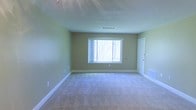 This single-floor space presents a straightforward and functional interior highlighted by a neutral color palette. The main room is finished with wall-to-wall carpet in a light beige tone, providing a soft underfoot feel throughout the space. The walls are painted in a muted green hue, lending a calm and understated ambiance to the interior.

A centrally placed window along the far wall allows natural light to filter into the room, offering a view of trees outside and giving the space an airy, connected-to-nature quality. White baseboards frame the perimeter of the room, providing a clean contrast against the green walls and light carpet.

The ceiling features a textured popcorn finish and includes a visible HVAC vent, indicating a functional climate control system integrated into the structure. A door on the right-hand wall provides access to an adjoining area or exit point, contributing to the overall flow of the layout.

The room is currently unfurnished, allowing for a clear assessment of its dimensions and features. The space appears well-proportioned, offering versatility for various uses. Overall, the interior maintains a clean, move-in-ready condition suitable for a range of configurations.