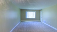 This single-floor space presents a straightforward and functional interior highlighted by a neutral color palette. The main room is finished with wall-to-wall carpet in a light beige tone, providing a soft underfoot feel throughout the space. The walls are painted in a muted green hue, lending a calm and understated ambiance to the interior.

A centrally placed window along the far wall allows natural light to filter into the room, offering a view of trees outside and giving the space an airy, connected-to-nature quality. White baseboards frame the perimeter of the room, providing a clean contrast against the green walls and light carpet.

The ceiling features a textured popcorn finish and includes a visible HVAC vent, indicating a functional climate control system integrated into the structure. A door on the right-hand wall provides access to an adjoining area or exit point, contributing to the overall flow of the layout.

The room is currently unfurnished, allowing for a clear assessment of its dimensions and features. The space appears well-proportioned, offering versatility for various uses. Overall, the interior maintains a clean, move-in-ready condition suitable for a range of configurations.