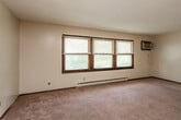 This single-floor unit offers a practical and well-organized layout across several distinct living spaces. The living area is generously sized, featuring neutral-toned carpet, a textured ceiling, a wall-mounted air conditioning unit, and a wide bank of three windows fitted with blinds that allow abundant natural light to fill the room. Baseboard heating runs along the perimeter, providing consistent warmth throughout. The kitchen is positioned adjacent to the living space and is equipped with dark wood cabinetry, a white electric range, a double-basin sink, and a full-size refrigerator. Stone-patterned vinyl flooring anchors the kitchen area, creating a clear visual distinction from the carpeted living zones. Two bedrooms are present, each featuring carpet flooring, textured ceilings, baseboard heating, and windows with blinds that offer both light and privacy. The rooms provide solid, functional spaces suited for a variety of configurations. The bathroom is compact and complete, featuring a tub-shower combination, a toilet, a single-sink vanity with under-cabinet storage, and yellow tile flooring. Dark wood trim and doors carry throughout the unit, lending a consistent and cohesive character to the overall interior aesthetic.