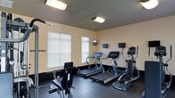 This fitness center is a dedicated, single-floor workout facility designed to meet a wide range of exercise needs. The space is outfitted with multiple treadmills and an elliptical machine, each equipped with individual digital display screens for tracking performance metrics. A strength training cable machine provides options for resistance-based workouts, rounding out the cardio-focused equipment lineup. The room features durable dark rubber flooring, ideal for high-impact activity and easy maintenance. Warm beige walls and bright fluorescent overhead lighting create a clean, energizing atmosphere. Natural light filters in through multiple windows fitted with white horizontal blinds, contributing to an open and airy feel. A wall-mounted mirror and flat-screen monitor are also present, adding both functional and entertainment value to the workout environment. The layout is thoughtfully organized to allow comfortable movement between equipment. This fitness center provides a practical and well-maintained space suited for daily exercise routines.