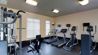 This fitness center is a dedicated, single-floor workout facility designed to meet a wide range of exercise needs. The space is outfitted with multiple treadmills and an elliptical machine, each equipped with individual digital display screens for tracking performance metrics. A strength training cable machine provides options for resistance-based workouts, rounding out the cardio-focused equipment lineup. The room features durable dark rubber flooring, ideal for high-impact activity and easy maintenance. Warm beige walls and bright fluorescent overhead lighting create a clean, energizing atmosphere. Natural light filters in through multiple windows fitted with white horizontal blinds, contributing to an open and airy feel. A wall-mounted mirror and flat-screen monitor are also present, adding both functional and entertainment value to the workout environment. The layout is thoughtfully organized to allow comfortable movement between equipment. This fitness center provides a practical and well-maintained space suited for daily exercise routines.