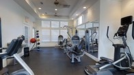 This fitness center is designed to support a comprehensive workout experience within a clean, well-appointed single-level layout. The space features durable rubber flooring throughout, providing a safe and functional surface for a wide range of exercises. Large windows with white blinds line the walls, allowing abundant natural light to fill the room and create an open, energizing atmosphere. A tray ceiling with recessed lighting and a ceiling fan adds architectural character while ensuring the space remains cool and well-lit during use. Strength training is well covered with multiple cable and resistance machines positioned throughout the floor, complemented by a wall-mounted mirror that visually expands the space and assists with form. A dedicated medicine ball rack along one wall offers a colorful array of weighted balls for functional fitness training. Additional cardio and strength equipment is visible throughout, rounding out the selection for a full-body workout. The overall ambiance is bright, clean, and purposefully arranged, making this fitness center a practical and inviting facility for daily exercise.
