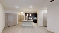 Located in Frisco, TX, this modern single-story space is designed with a clean, contemporary aesthetic that emphasizes open living and functional design. The heart of the space is an open-concept kitchen that flows seamlessly into the main living area. A large central island with a granite countertop serves as both a prep space and a gathering point, illuminated by stylish pendant lights hanging from the ceiling. The kitchen is outfitted with rich, dark espresso cabinetry that provides a striking contrast against the light-toned walls and backsplash. Stainless steel appliances, including a refrigerator, range, and microwave, complete the kitchen's polished, modern look. Recessed lighting and track lighting throughout ensure the space is well-lit and versatile. Light wood-look plank flooring runs throughout the main area, lending warmth and continuity to the design. Multiple doorways visible from the main space lead to additional rooms, suggesting a well-organized and practical layout. The overall ambiance is bright, airy, and move-in ready, reflecting the upscale residential character typical of the Frisco, TX area.