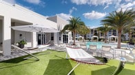 This outdoor amenity space exudes a vibrant, resort-inspired atmosphere designed for relaxation and leisure. At its center sits a large, crystal-clear swimming pool, flanked by rows of white lounge chairs that invite sunbathing and unwinding. The surrounding deck seamlessly transitions to a well-manicured artificial grass lawn, where freestanding hammocks offer a casual and comfortable retreat. Oversized cantilever umbrellas provide shaded respite throughout the space, ensuring comfort on sunny days. Tall, mature palm trees are strategically placed throughout the area, enhancing the tropical ambiance while adding natural vertical interest against the bright blue sky. The adjacent single-story building features clean, modern white architecture with large glass French doors that connect the indoor and outdoor spaces, allowing for easy flow between the two. Additional lounge areas and cabana-style seating can be seen extending further along the pool deck in the background, suggesting ample space for gatherings. The overall aesthetic is bright, airy, and polished, reflecting a contemporary Florida-inspired design sensibility well-suited for an active, social lifestyle.