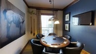 This single-floor space is designed as a dedicated game and recreation room, offering a comfortable and inviting atmosphere for social entertainment. The centerpiece of the room is a large, round wooden poker and game table, complete with built-in cup holders and playing surface details, surrounded by plush black leather club chairs that provide comfortable seating for multiple guests.

Overhead, a wrought iron chandelier with warm lamp shades casts ambient light directly over the table, enhancing the room's intimate feel. A window dressed with plaid curtains and horizontal blinds allows natural light to filter in, brightening the space during daytime use.

The décor leans into a rustic, lodge-inspired aesthetic. Decorative wooden oars and a vintage model airplane are mounted on the walls, while a large-scale animal artwork anchors one side of the room. The navy blue accent wall on the opposite side is adorned with a series of framed botanical and nature prints, adding visual interest and depth.

Crown molding with dark wood trim borders the ceiling, lending architectural refinement. The carpeted floor and neutral wall tones create a warm, grounded ambiance throughout the space.