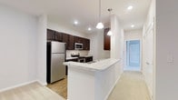 This single-floor space showcases a well-designed open-concept kitchen as its centerpiece. The kitchen is outfitted with rich, dark espresso cabinetry that extends to the upper walls, providing ample storage while creating a bold visual contrast against the crisp white walls and light-toned flooring. A suite of stainless steel appliances — including a full-size refrigerator, microwave, and gas range — adds a contemporary, functional edge to the space.

The kitchen peninsula features a light granite countertop with subtle veining, offering additional prep space and serving as a natural divider between the kitchen and the adjacent living area. Two pendant lights hang above the peninsula, providing focused task lighting that complements the recessed ceiling lights throughout.

The ceilings are notably tall, enhancing the open and airy feel of the space. The flooring transitions from hard surface in the kitchen area to carpet in the adjoining room, adding warmth and texture. A hallway visible beyond the kitchen leads to additional rooms, suggesting a thoughtfully connected single-level layout. Overall, the space presents a clean, modern aesthetic with practical design choices throughout.