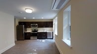 This single-floor space centers around a well-appointed kitchen that serves as the focal point of the open layout. The kitchen features rich, dark espresso-toned cabinetry extending along the full wall, providing ample storage through both upper and lower cabinets. Stainless steel appliances, including a refrigerator, electric range, built-in microwave, and dishwasher, complement the cabinetry and contribute to a cohesive, modern aesthetic. A clean, light-toned countertop runs the length of the kitchen, offering generous workspace, while a stainless steel sink is positioned beneath natural light filtering through roller-shaded windows. Recessed lighting supplements the flush-mount ceiling fixture, ensuring the space is well-lit throughout. A wall-mounted HVAC unit is integrated into the ceiling, providing efficient climate control. The flooring throughout is a smooth, light-toned hard surface that reflects light and enhances the sense of openness. Large windows along the adjacent wall flood the space with natural light, creating a bright, airy ambiance. The overall design balances functionality with a clean, contemporary style.