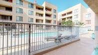The pool area at 606 3rd Street in San Diego is a well-appointed outdoor amenity set within a landscaped courtyard framed by multi-story residential buildings. The space features a generously sized swimming pool with clear, blue-green water, accessible via stainless steel entry rails. The surrounding deck is finished in warm terracotta-toned tile and hexagonal pavers, providing ample space for sunbathing and relaxation.

Multiple chaise lounges are positioned along the poolside for individual use, while a shaded dining area with umbrella-covered tables and chairs offers a comfortable spot for outdoor gatherings. The courtyard is enclosed by metal rail fencing with a gated entry point, ensuring a secure and defined pool environment.

Architecturally, the surrounding buildings feature a warm sandy stucco exterior with arched entryways, teal tile accents, and pink-hued columns that lend a distinctive Southwestern aesthetic to the space. Lush planters with ground cover greenery add a touch of natural softness alongside the hardscape.

The pool facility includes a dedicated entry vestibule with glass doors, hexagonal tile flooring, and arched detailing, creating a welcoming transition point. The open courtyard orientation allows for abundant natural sunlight throughout the day, enhancing the overall outdoor experience in this San Diego setting.