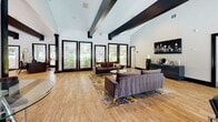 Located at 2005 Treehills Pkwy in Stone Mountain, Georgia, this single-floor space delivers a polished, contemporary environment designed for comfort and functionality. The interior is defined by dramatically vaulted ceilings with bold dark exposed beams that contrast sharply against crisp white walls, creating a striking architectural focal point throughout the open layout.

Light wood flooring runs continuously across the space, contributing warmth and cohesion. A generous wall of large, black-framed windows lines one side of the room, flooding the interior with abundant natural light and offering views of the surrounding greenery. The black trim and framing details on doorways and windows provide consistent, modern contrast throughout.

The main gathering area features plush velvet sofas in deep charcoal-purple tones, accented with gold-legged seating and layered area rugs in abstract gold and gray patterns. A separate seating zone incorporates sleek chrome and leather chairs paired with glass-topped bistro tables, offering a more structured setting. Contemporary wall art and a city-themed canvas add visual interest, while a beverage station is tucked neatly along one wall. Additional private rooms are accessible through bold black-framed doorways, rounding out this well-appointed, design-forward space.