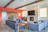 Located at 633 W Main St in Sun Prairie, Wisconsin, this single-floor community clubhouse offers a generously proportioned, amenity-rich gathering space designed with modern comfort and style in mind. Exposed wooden beam ceilings and light wood-look tile flooring run throughout, creating a warm, cohesive aesthetic.

The main lounge area serves as the social heart of the space, anchored by a striking floor-to-ceiling stacked stone fireplace with a mounted flat-screen TV above. Bold terracotta-orange accent walls and sliding barn doors add character, while plush sectional sofas and lounge chairs arranged around the fireplace invite relaxed conversation. Pendant lighting and a chandelier overhead complement the vaulted beam ceiling above.

The fully equipped kitchen features dark wood cabinetry, granite countertops, stainless steel appliances including a large refrigerator and gas range, a herringbone tile backsplash, and open shelving. A long granite-topped island with bar seating accommodates group gatherings with ease.

An adjoining dining area is bathed in natural light from a continuous bank of oversized windows overlooking an outdoor pool and patio, offering a bright and airy atmosphere ideal for casual dining or working. A dedicated theater room rounds out the space, complete with tiered seating, ambient underlighting, a projector, and classic movie poster décor.