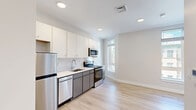 Located at 72 Burbank Street, Unit 405, in Boston's Fenway neighborhood, this efficiently designed unit offers a thoughtful layout across a single floor. The open living area is bathed in natural light through multiple windows overlooking the surrounding streetscape, with light-toned hardwood flooring and crisp white walls creating a clean, contemporary atmosphere throughout.

The kitchen is well-appointed with two-tone cabinetry — white upper cabinets paired with gray lower cabinets — and features a full suite of stainless steel appliances, including a refrigerator, gas range, built-in microwave, and dishwasher. A white countertop with an undermount sink completes the functional workspace, all set atop the same light hardwood floors that carry through the living area.

The bathroom showcases a refined aesthetic with striking marble-style porcelain tile flooring and a frameless glass walk-in shower with white subway tile surround. A floating-style vanity with a white countertop and gray cabinetry ties together the modern design. Recessed lighting and a neutral white palette lend the bathroom a bright, spa-like quality. A smart home panel near the entryway adds a modern, connected touch to this polished Boston residence.